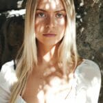 A blond woman in a white top leaning against a stone wall.
