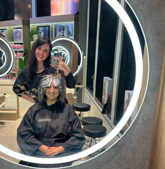 Two women taking a selfie in front of a mirror in a salon, while one woman adjusts her Aline haircut.