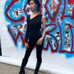 A woman in a black jumpsuit standing in front of a graffiti wall.
