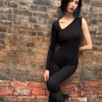 A woman in a black jumpsuit leaning against a brick wall.