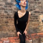 A woman with blue hair leaning against a brick wall.