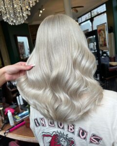 Person with long, wavy, platinum blonde hair being styled in a salon, wearing a t-shirt with "Incubus" printed on it.