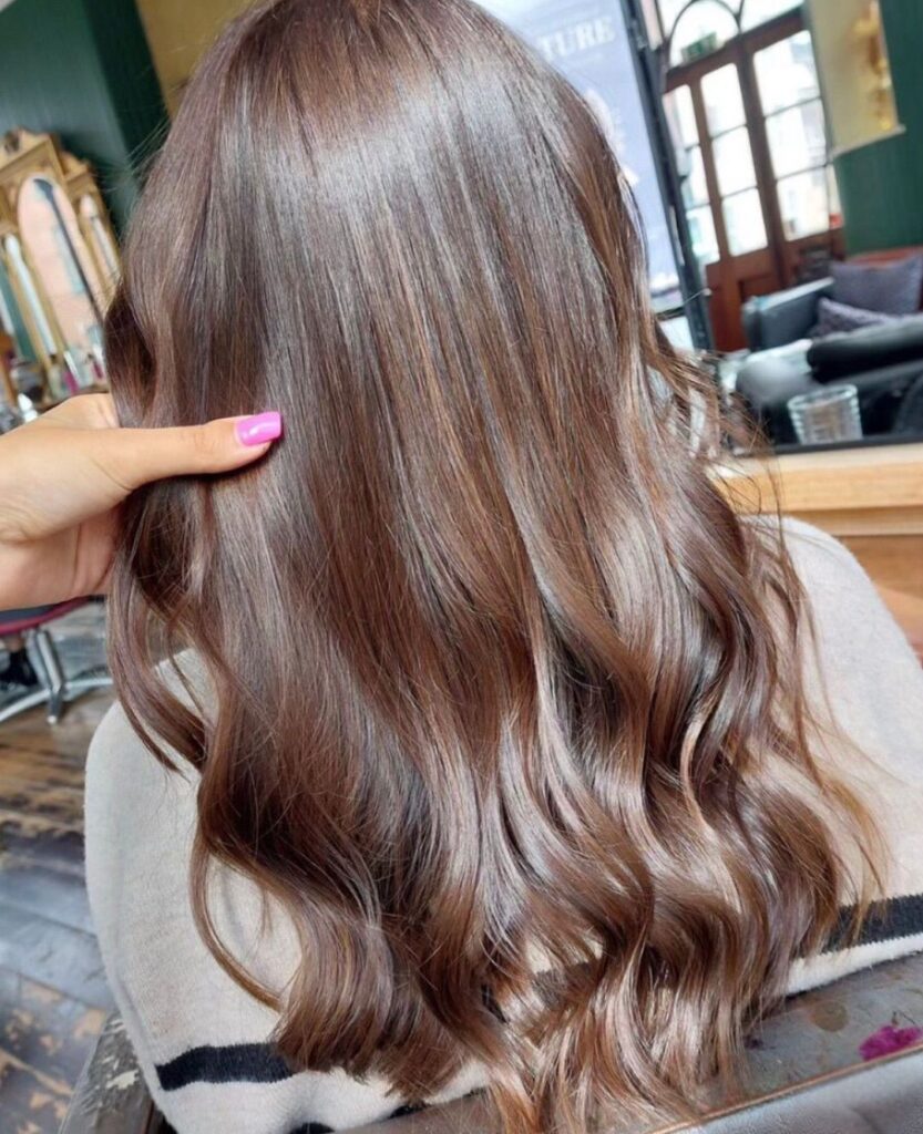 A person with long, wavy, light brown hair sits in a salon. A hand with pink nails is pointing at the hair. Mirrors and salon furniture are visible in the background.