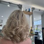 A person with shoulder-length, wavy blonde hair is seated in front of salon mirrors at Stream Salon, with shelves and products visible in the background.
