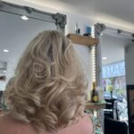 A woman with shoulder-length, wavy blonde hair sits in front of a stream salon mirror, surrounded by stylish hair products and decor in the background.