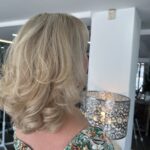 A person with shoulder-length, wavy blonde hair—fresh from the stream salon—wearing a floral-patterned top, is facing away from the camera near a lit decorative lamp.