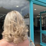 A person with shoulder-length, wavy blonde hair stands outside Stream Salon, its turquoise trim framing the entrance, facing away from the camera.