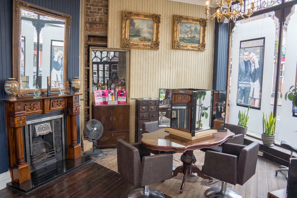 With its vintage charm, our hair salon in Portsmouth features wooden flooring, a decorative fireplace, mirrors, and framed art. Two styling chairs encircle a small round table. Large windows display promotional posters affirming our status as one of the top 3 salons near me in portsmouth.