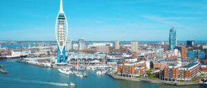 A waterfront cityscape with a prominent white tower, modern and historic buildings, and boats docked in the harbor under a clear blue sky, mirrors the charm of Portsmouth's top salons nestled nearby.