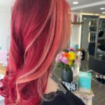 Person with vibrant red and pink streaked hair styled in loose waves, in a salon setting with flowers and styling products in the background.