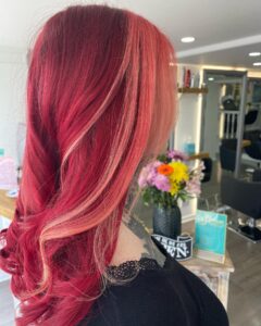 Person with wavy, bright red and pink highlighted hair, wearing a black top, in a salon setting with flowers and salon products in the background.