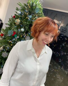 A woman with short red hair and a white blouse smiles while standing in front of a decorated Christmas tree with lights and ornaments.