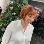 A person with red hair stands in front of a decorated Christmas tree, wearing a white shirt and smiling.