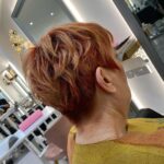 Person with short, red hair seated in a salon chair, facing away. The background shows salon mirrors and equipment.