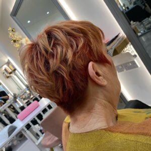 Person with short, red hair seated in a salon chair, facing away. The background shows salon mirrors and equipment.