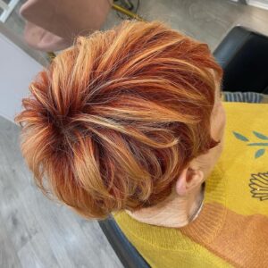 A person with short, voluminous hair dyed in shades of red and blonde sits in a salon chair, wearing a yellow sweater.