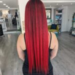 Person with long, straight red hair stands in a salon, facing away. They wear a black sleeveless top. The salon has wooden flooring and modern decor.