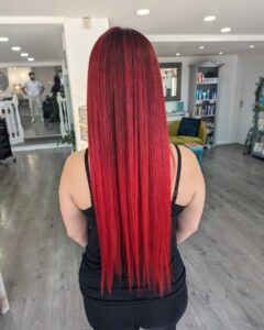 Person with long, straight red hair stands in a salon, facing away. They wear a black sleeveless top. The salon has wooden flooring and modern decor.