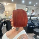Person with a red bob haircut sits in a salon chair. Tattoo visible on shoulder. Background shows a stylist and hair care products.