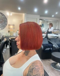 Person with a red bob haircut sits in a salon chair. Tattoo visible on shoulder. Background shows a stylist and hair care products.