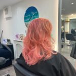 Person with wavy, shoulder-length pink hair sitting in a salon chair, facing away. White walls and a beach-themed wall art visible.