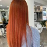 Person with straight, long, reddish-orange hair stands in a salon, facing away. They wear a light blue sweater.