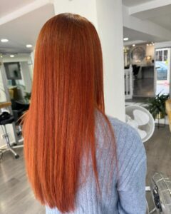 Person with long, straight, vibrant red hair wearing a blue sweater, standing indoors in a salon with white walls and modern decor.
