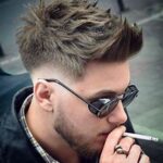 A man with spiked hair, wearing sunglasses and a checked shirt, smokes a cigarette while sitting outdoors.