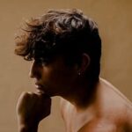 A young person with short, wavy brown hair sits shirtless in profile against a plain beige background, resting their chin on their hand.
