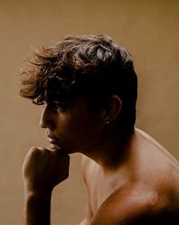 A shirtless person with short, curly brown hair sits in profile against a neutral background, resting their chin on their hand.