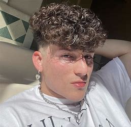 Young person with curly brown hair, wearing a white t-shirt, chain necklace, and heart-shaped earring, reclines in sunlight with their hand behind their head.