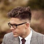 A man wearing glasses, a gray plaid suit, white shirt, and maroon tie, with styled short hair, looking to the side outdoors.