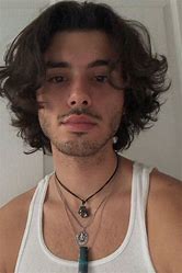 A young man with medium-length wavy hair wearing a white tank top and layered necklaces stands in front of a white background.