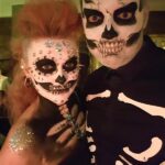 Two people wearing skeleton makeup and costumes, with one person also decorated with rhinestones and glitter on their face, chest, and shoulder.