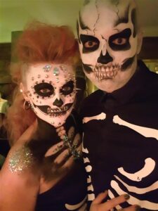 Two people wearing skeleton makeup and costumes, with one person also decorated with rhinestones and glitter on their face, chest, and shoulder.