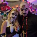 Two people in occult-themed costumes with pentagram face paint pose in front of a wall decorated with colorful sugar skulls and string lights.