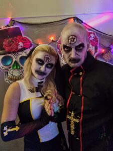 Two people in occult-themed costumes with pentagram face paint pose in front of a wall decorated with colorful sugar skulls and string lights.