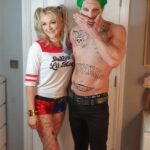 Two people in costume stand side by side; one is dressed as Harley Quinn and the other as the Joker, both from DC Comics, posing indoors in front of a closed door.