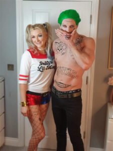 Two people in costume stand side by side; one is dressed as Harley Quinn and the other as the Joker, both from DC Comics, posing indoors in front of a closed door.