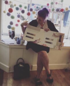 A person with purple hair sits by a window, holding and pointing to a color sample book, with colorful pom-poms hanging in the background.