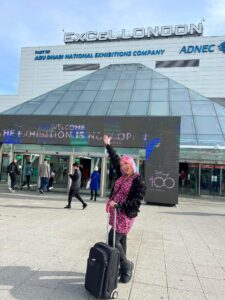 Person with pink hair and a suitcase stands outside ExCeL London entrance, pointing up at the venue sign; other people walk in and out of the building.