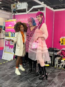 Three people pose and smile together at a convention booth with a pink backdrop and promotional signs, wearing colorful and distinctive outfits.
