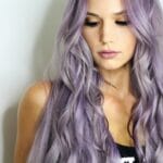 A woman with long purple hair is posing in front of a white wall.