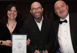 Three smiling people in formal attire are holding awards. The woman on the left holds a certificate, while the man in the middle and the man on the right each hold a trophy.