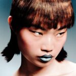 A person with short, geometric brown hair, straight bangs, and metallic blue lipstick looks to the side with a serious expression against a neutral background.