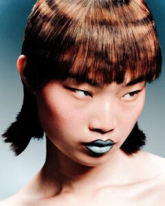 A person with short, geometric brown hair, straight bangs, and metallic blue lipstick looks to the side with a serious expression against a neutral background.