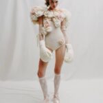 A person stands against a white backdrop wearing a floral bodysuit, lace socks, satin pointe shoes, and oversized fabric boxing gloves.
