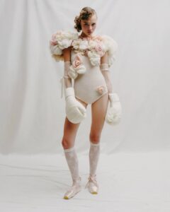 A person stands against a white backdrop wearing a floral bodysuit, lace socks, satin pointe shoes, and oversized fabric boxing gloves.