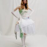 Person wearing a layered white dress with lace gloves, knee-high lace socks, and a green sash that reads “Dream.” Neutral backdrop.