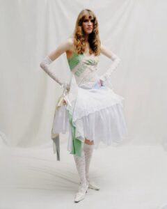 Person wearing a layered white dress with lace gloves, knee-high lace socks, and a green sash that reads “Dream.” Neutral backdrop.